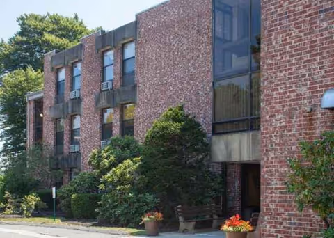 Brick senior living facility exterior with trees, planters, and a visible entrance.
