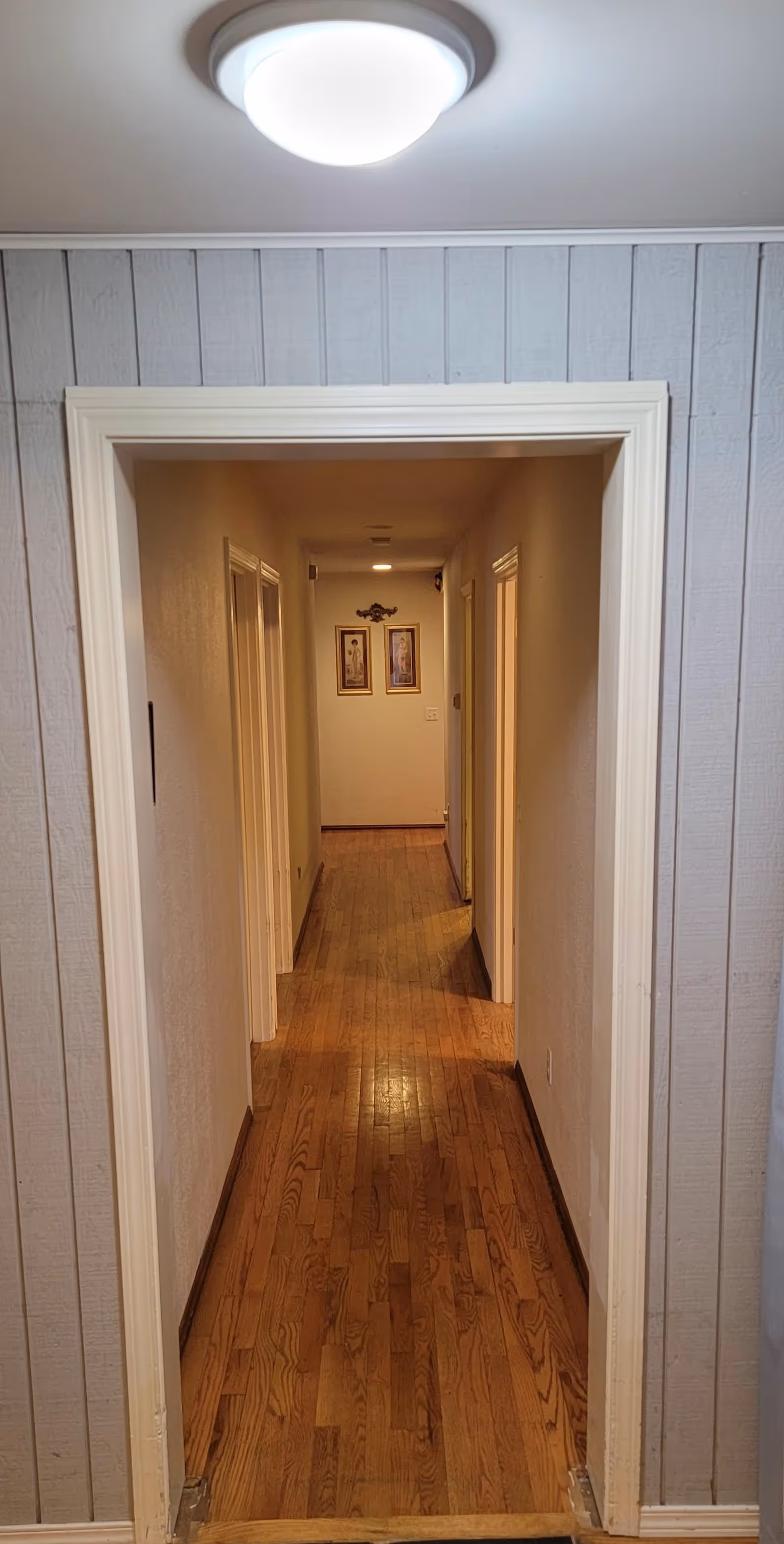 A long narrow interior hallway with hardwood floors, doorways on both sides, and framed pictures at the far end.