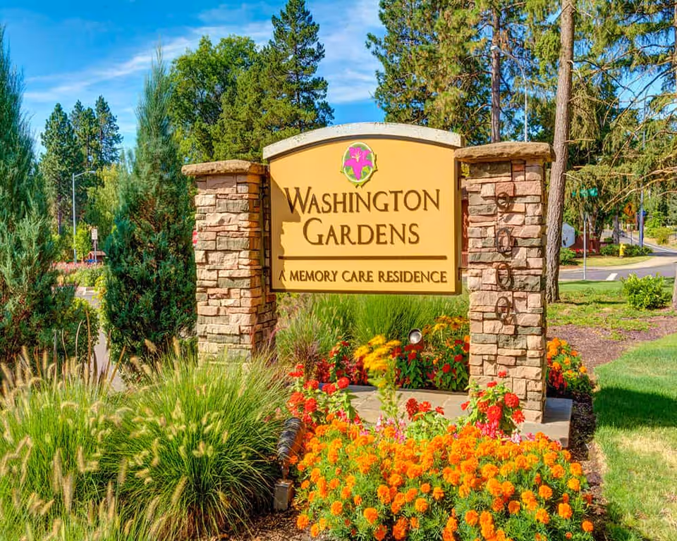 Outdoor stone sign for Washington Gardens Memory Care Residence surrounded by colorful flowers and greenery with trees and a road in the background under a blue sky.