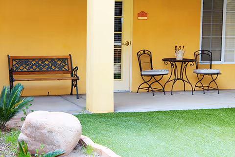 Outdoor patio area with a yellow wall background featuring a wooden bench with metal armrests on the left, a small round metal table with two cushioned metal chairs on the right, a door with a window in the center, and a large rock with some plants in the foreground on a grassy lawn.