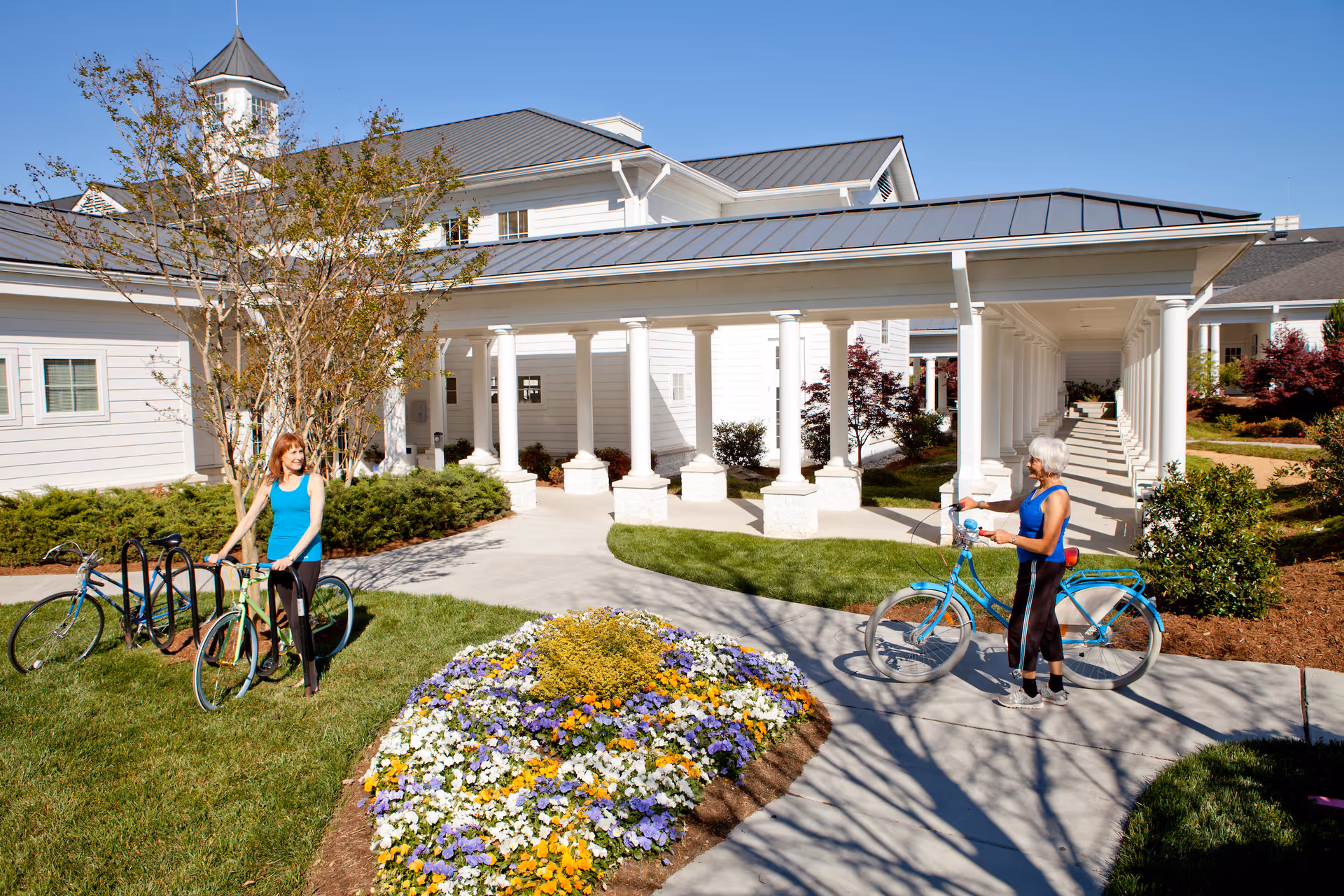 Two women with bicycles stand on a pathway in front of the columned entrance of a white senior living building with flowerbeds.