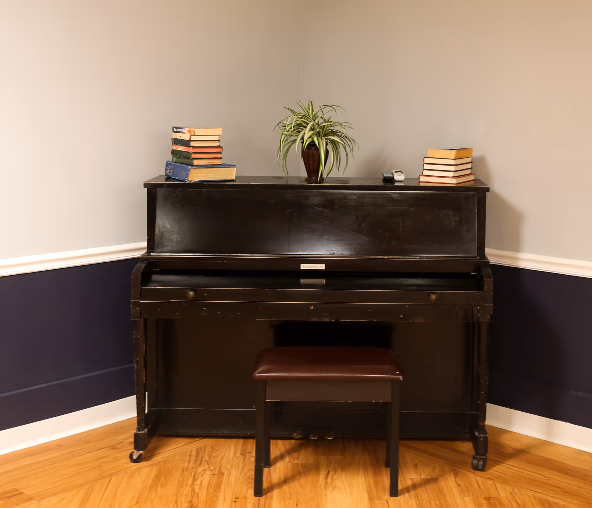 A black upright piano with a matching bench in front of it, placed against a two-tone wall with beige on top and dark blue on the bottom. On top of the piano are two stacks of books on either side and a potted plant in the center.