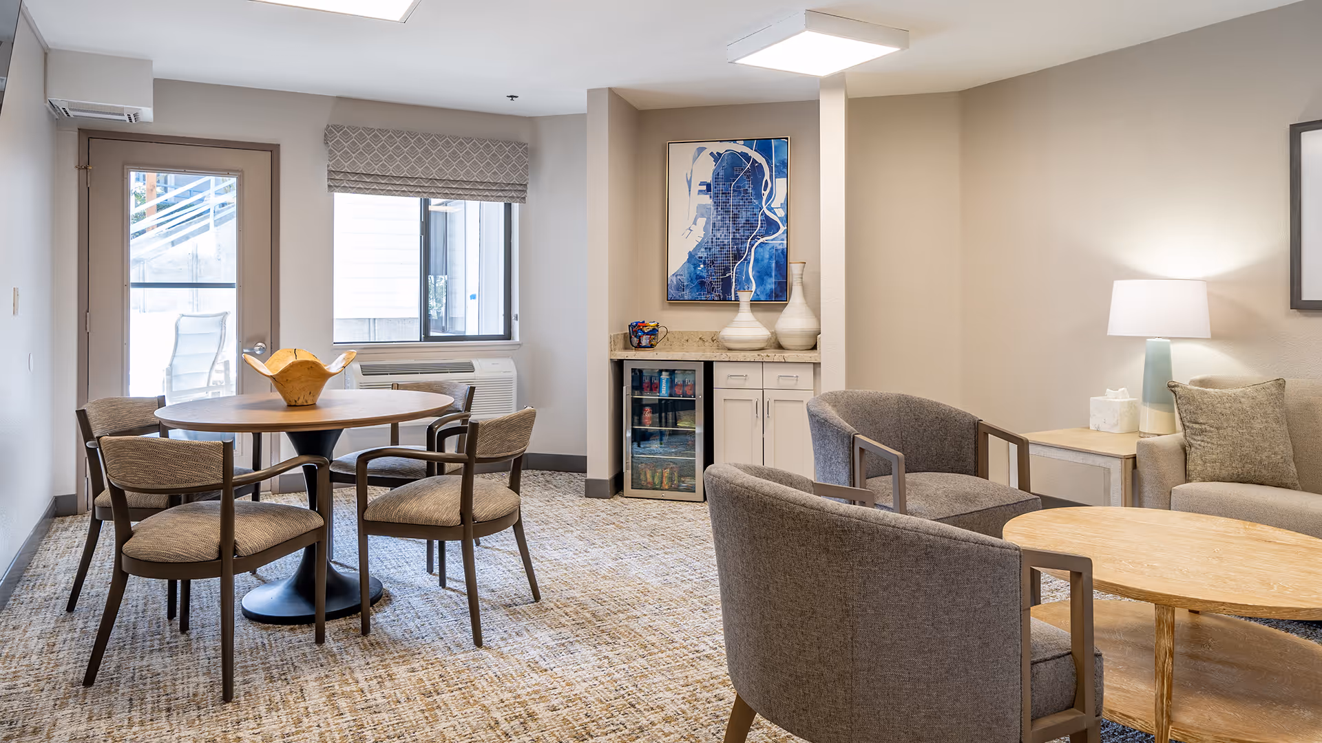 A bright and cozy common area in a senior living facility featuring a round wooden table with four chairs, a small sitting area with two gray armchairs and a wooden coffee table, a beige sofa with cushions, a side table with a lamp and tissue box, a small cabinet with a mini fridge, and a framed abstract blue artwork on the wall. There is a door and window letting in natural light.