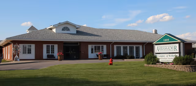 Single-story brick building with a gray shingled roof under a blue sky with some clouds. There is a green lawn in front and a sign that reads 'Ambassador Campus of Wellness' near the entrance.