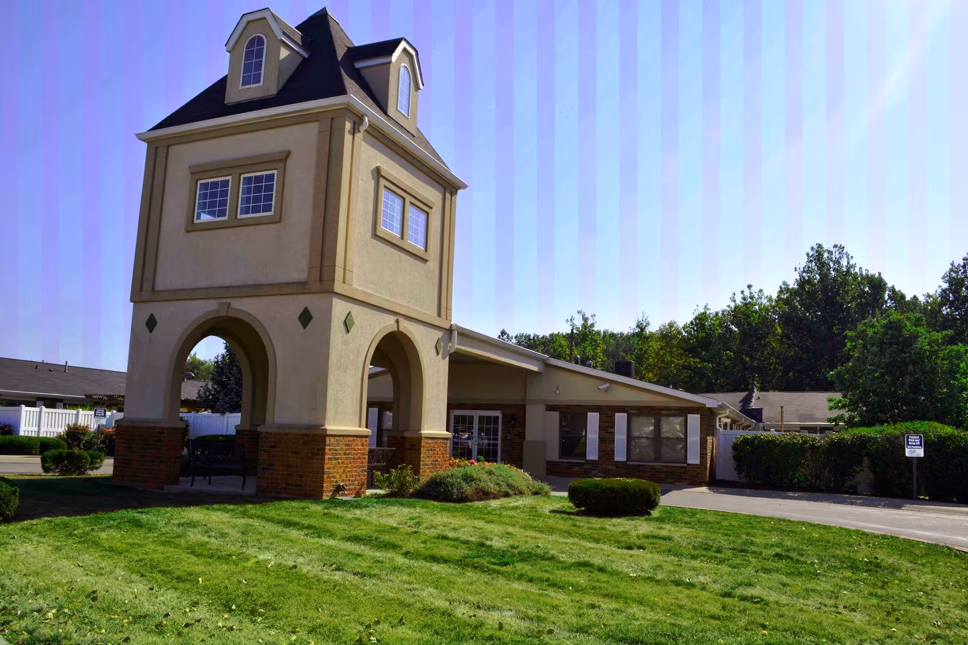 Exterior view of a senior living facility named Riverwalk Village featuring a unique tower-like structure with arched openings and windows, surrounded by a well-maintained lawn and greenery under a clear blue sky.