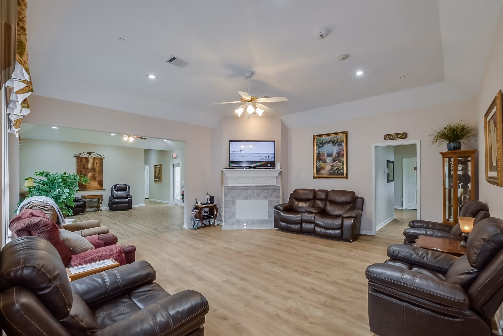 A spacious living room with multiple brown leather recliners and a couple of fabric armchairs arranged around a fireplace with a mounted flat-screen TV above it. The room has light-colored walls, wood flooring, ceiling fan with lights, and decorative paintings on the walls. There is an open doorway leading to another seating area with more chairs and a large green plant.