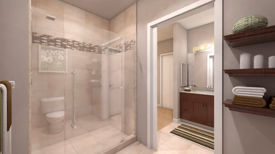 A modern bathroom featuring a glass-enclosed shower with beige tiles and a decorative tile border. Inside the shower area, a toilet is visible. To the right, there is a vanity with a sink, a mirror, and a light fixture above. Wooden shelves on the right hold neatly folded towels and decorative items. The bathroom opens into a carpeted hallway.