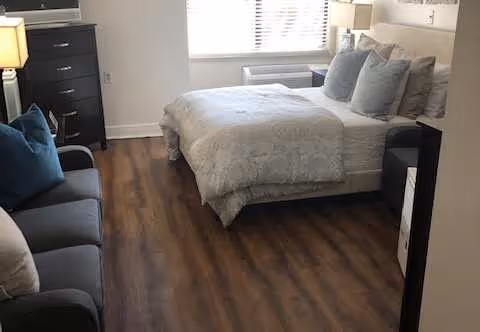Small furnished bedroom with a bed, sofa, dresser, lamp, and wood-look flooring under a window.
