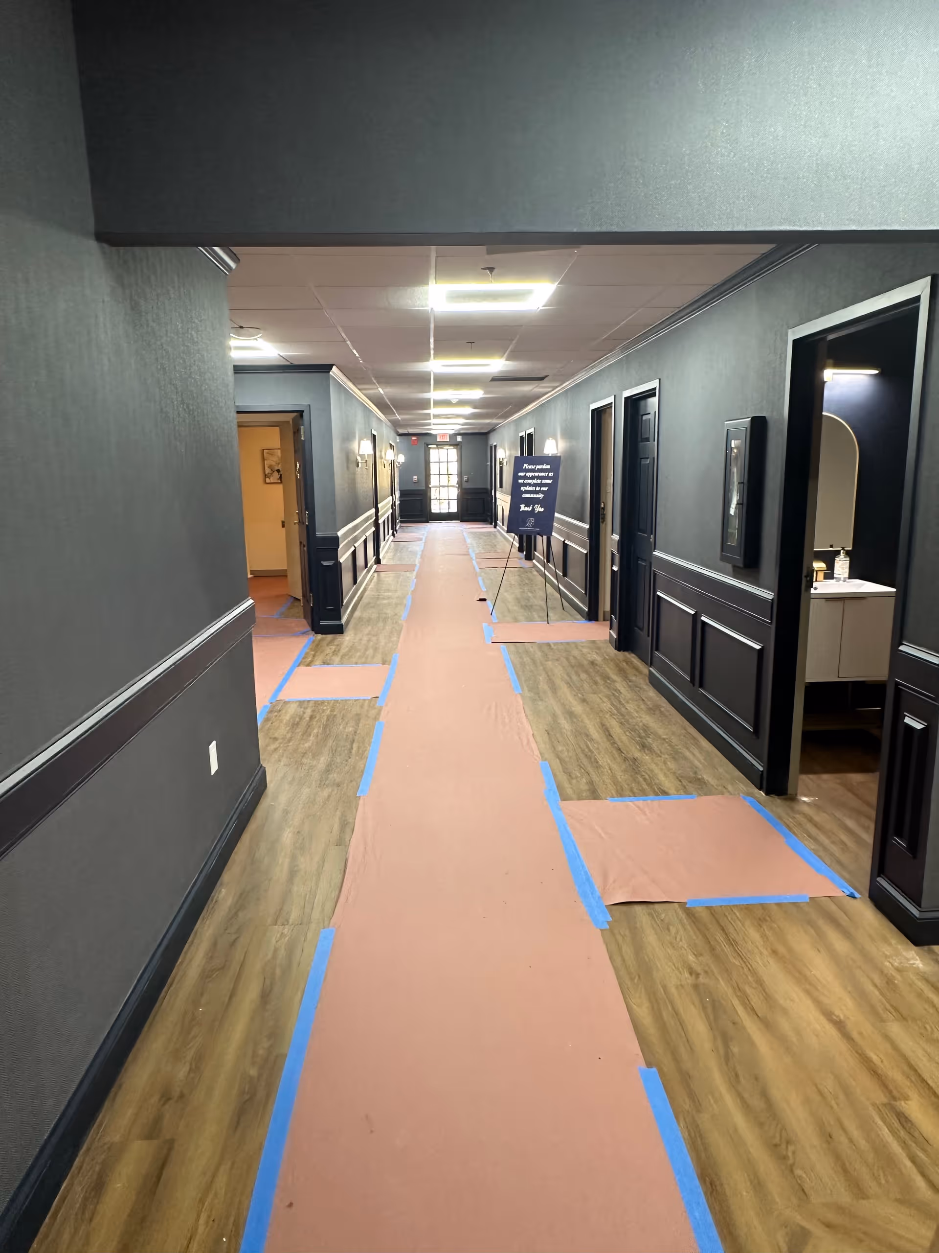 A long hallway in a senior living facility with dark gray walls and wood flooring partially covered by protective paper taped down with blue tape. Several doorways line the hallway, one showing a sink and mirror inside. A sign is placed on a stand further down the hallway near an exit door with glass panels.