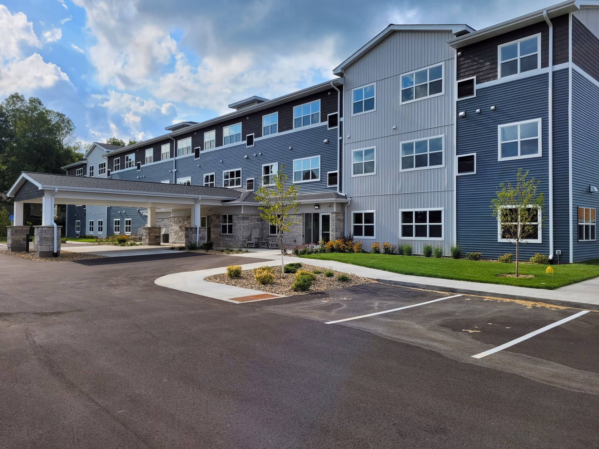 Exterior view of a modern three-story senior living facility with blue and gray siding, multiple windows, a covered entrance, small landscaped areas with young trees and shrubs, and an empty parking lot under a partly cloudy sky.