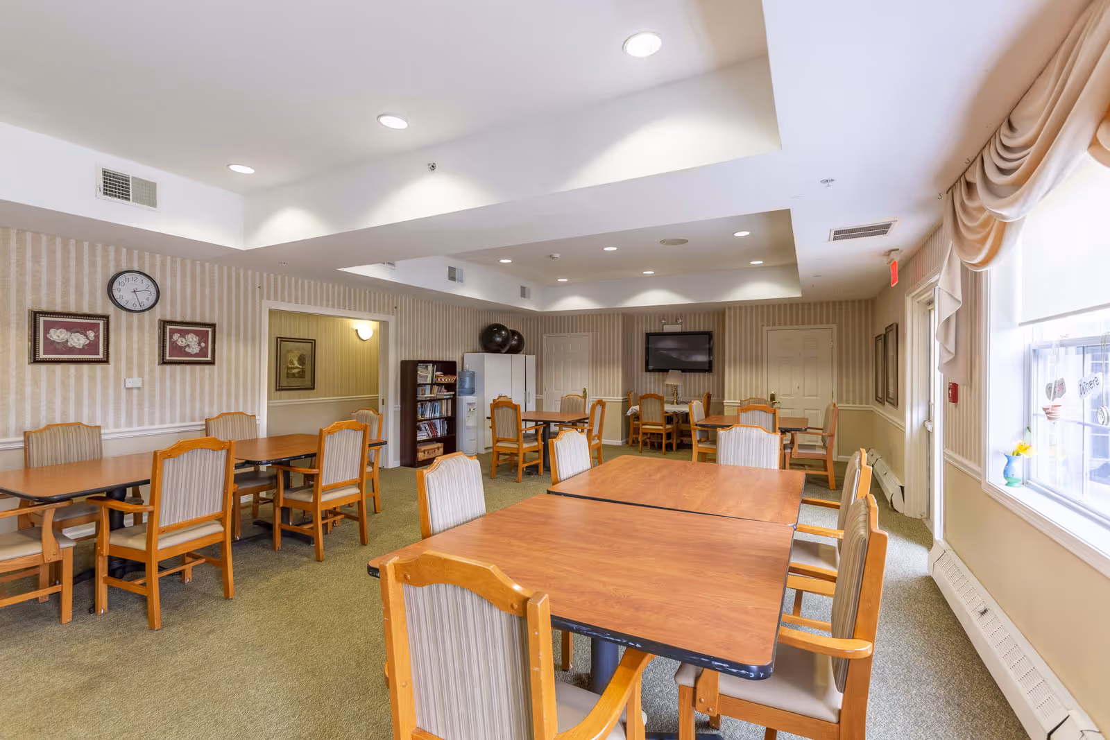 A well-lit dining room with multiple wooden tables and cushioned chairs arranged neatly. The room has beige striped wallpaper, a green carpet, and recessed ceiling lights. There is a clock and framed floral artwork on one wall, a bookshelf with books and a water dispenser, and a flat-screen TV mounted on another wall. A window with draped curtains allows natural light to enter the room.