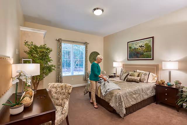 A senior woman making the bed in a well-lit bedroom with beige walls and carpet. The room features a large window with patterned curtains, a wooden desk with a chair, a potted plant, bedside tables with lamps, and a framed painting above the bed.