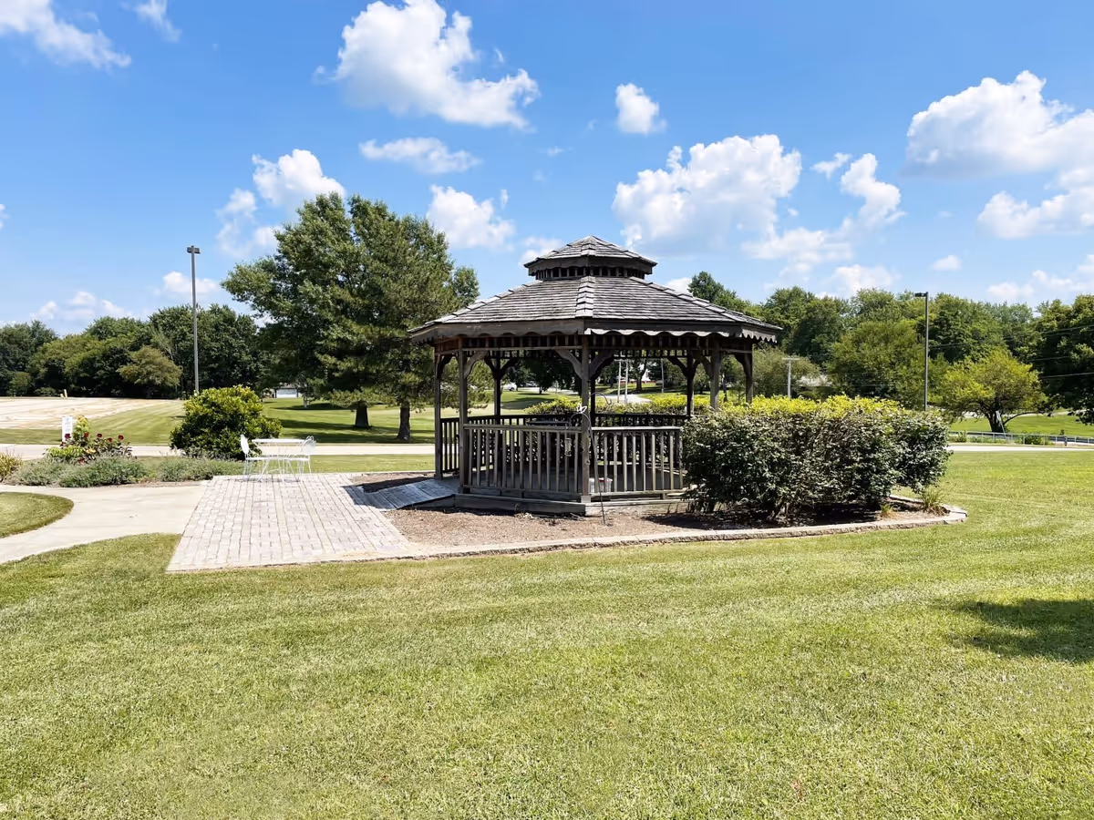 A wooden gazebo with a shingled roof situated on a grassy lawn under a blue sky with scattered clouds. There are bushes around the gazebo and a paved walkway leading to a white metal table and chairs set on the left side. Trees and a parking lot are visible in the background.