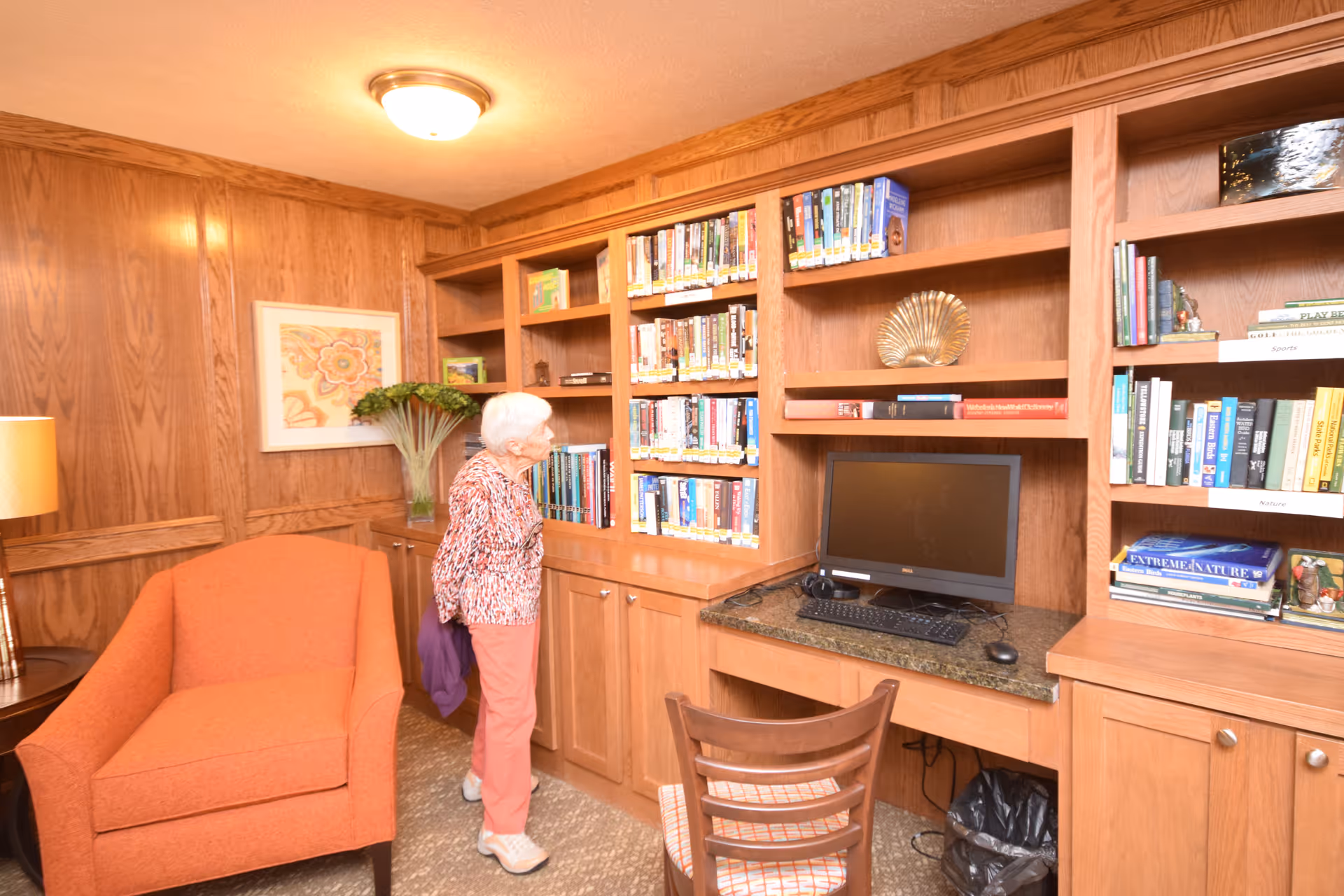 An elderly woman stands in a cozy room with wood-paneled walls, looking at a bookshelf filled with books. The room features an orange armchair, a wooden desk with a computer monitor, keyboard, and mouse, and a wooden chair with a patterned cushion. A framed floral artwork and a vase with green plants are on a side table next to the armchair.
