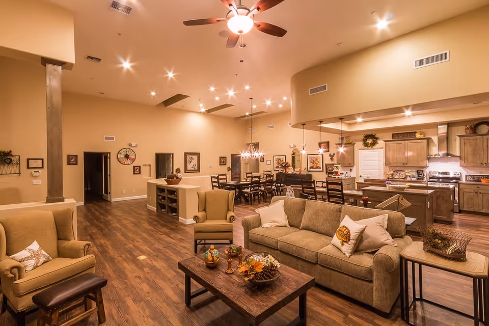 Spacious and warmly lit common area featuring a living room with beige sofas and armchairs, a wooden coffee table with decorative items, and a dining area with a large wooden table and chairs. The background shows a kitchen with wooden cabinets, stainless steel appliances, and pendant lighting. The room has wooden flooring and beige walls with various framed artworks and decorations.