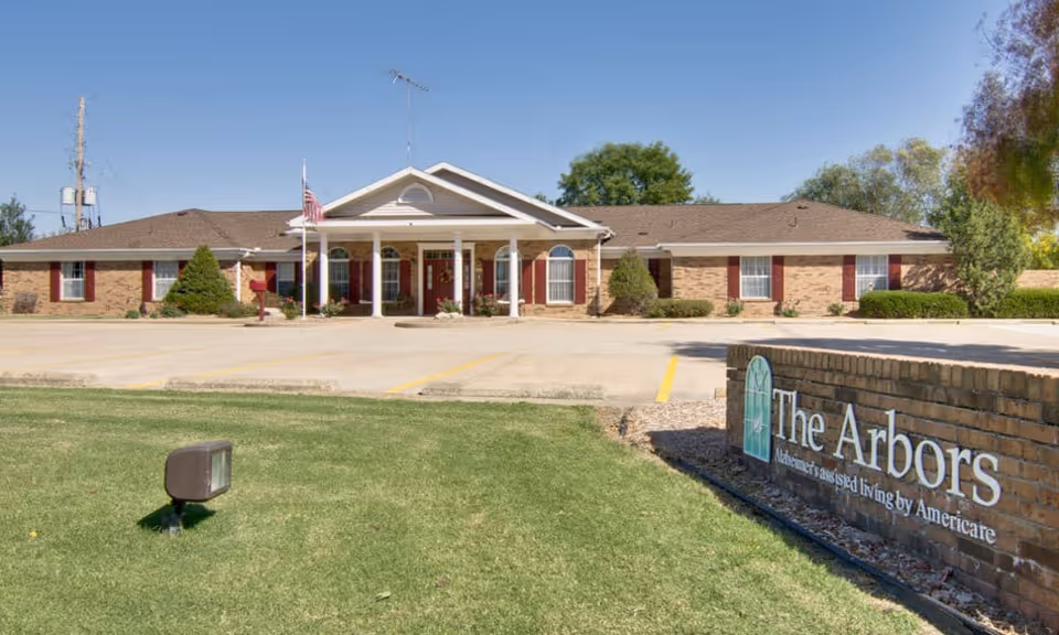 Front exterior of a single-story brick senior living facility with a columned entrance, flagpole, and a 'The Arbors' sign on a brick wall.