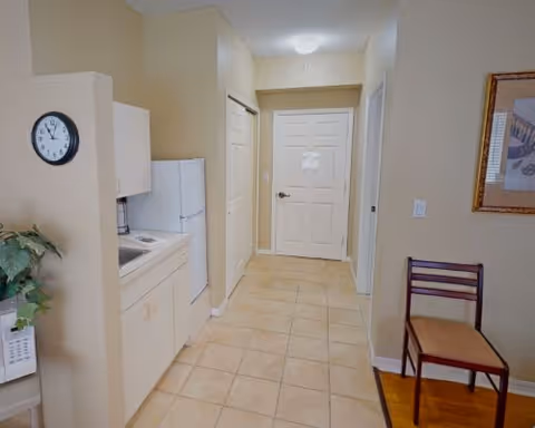 A small kitchen area with a white refrigerator, white cabinets, a microwave, and a clock on the wall. The kitchen opens into a hallway with a closed white door at the end. A wooden chair with a cushion is placed against the right wall, which also has a framed picture hanging above it. The floor is tiled in the kitchen and hallway area, with a transition to wood flooring near the chair.