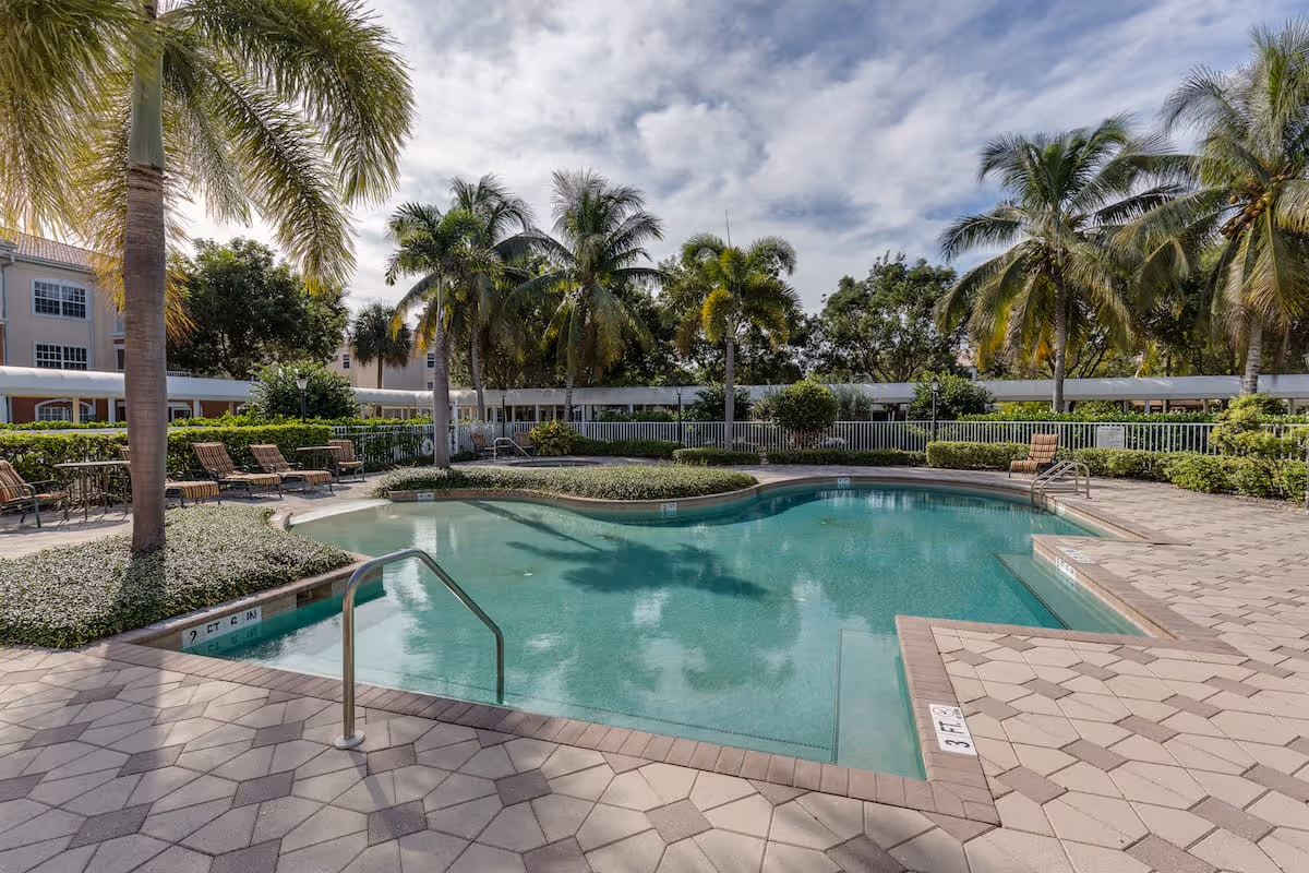 Outdoor swimming pool surrounded by palm trees and lounge chairs at Aston Gardens At Pelican Marsh, with a partly cloudy sky overhead.