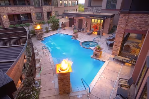 Outdoor swimming pool area in a residential or senior living facility with fire features on stone pillars, lounge chairs, a hot tub, and water fountains spraying into the pool, surrounded by multi-story buildings with balconies.