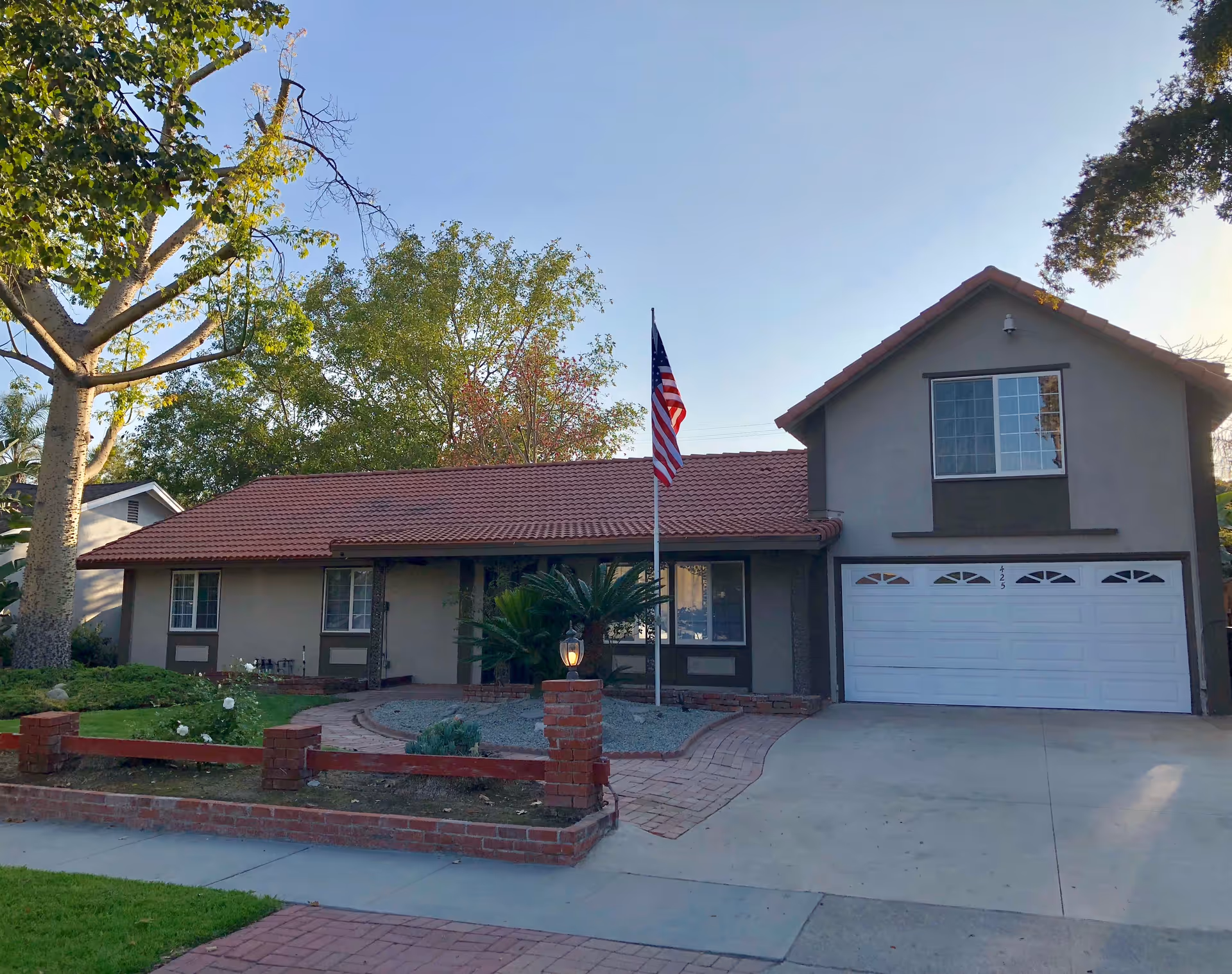 Front exterior view of a single-story residential building with a two-car garage, a red-tiled roof, and an American flag on a flagpole in the front yard. The yard has a brick-lined pathway, some greenery, and trees surrounding the house.