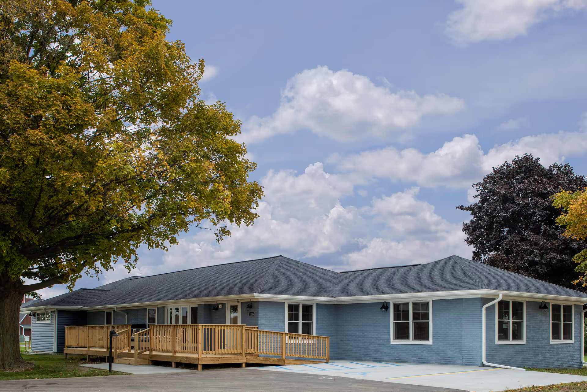 A blue senior living facility with a wooden ramp and large windows, surrounded by trees and a cloudy sky.