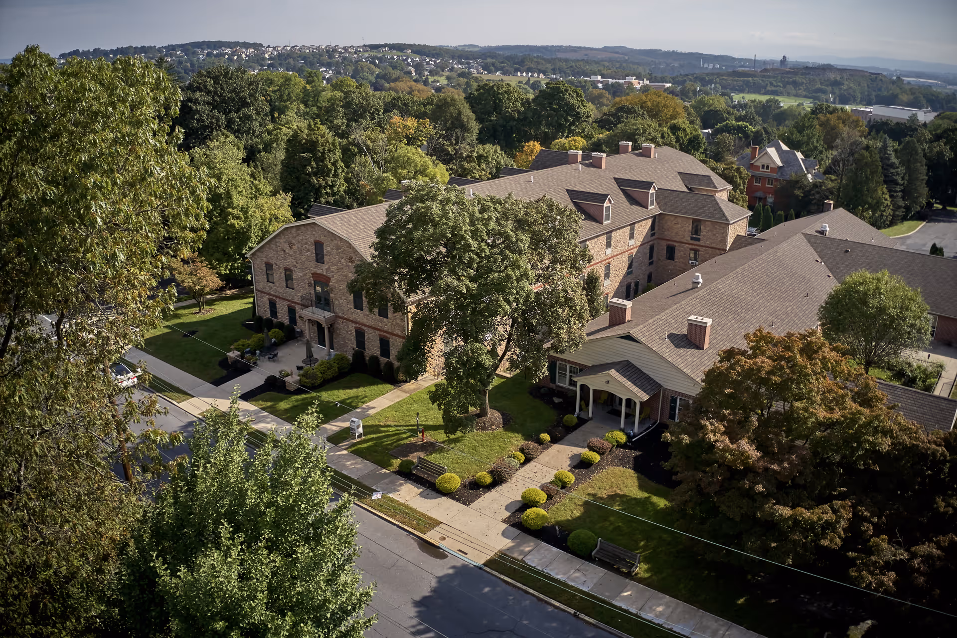 Aerial view of Alexandria Manor, a senior living facility surrounded by trees and greenery. The building is made of brick with multiple chimneys and a well-maintained lawn with shrubs and benches. The surrounding area includes roads and more trees extending into the distance under a clear sky.
