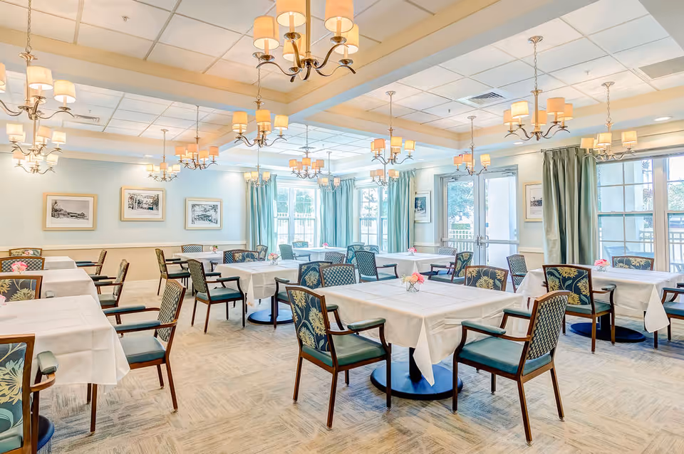 A bright and spacious dining room with multiple tables covered in white tablecloths, surrounded by chairs with floral and patterned upholstery. The room features large windows with green curtains, several chandeliers with multiple lampshades hanging from the ceiling, framed artwork on the walls, and a door leading outside.