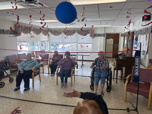 A group of elderly women seated in chairs and wheelchairs inside a decorated room with hanging streamers and a large blue balloon. They are positioned behind a badminton net, appearing to be engaged in a seated game or activity. The room has large windows with blinds and patterned valances, and there is a piano and bulletin board visible in the background.