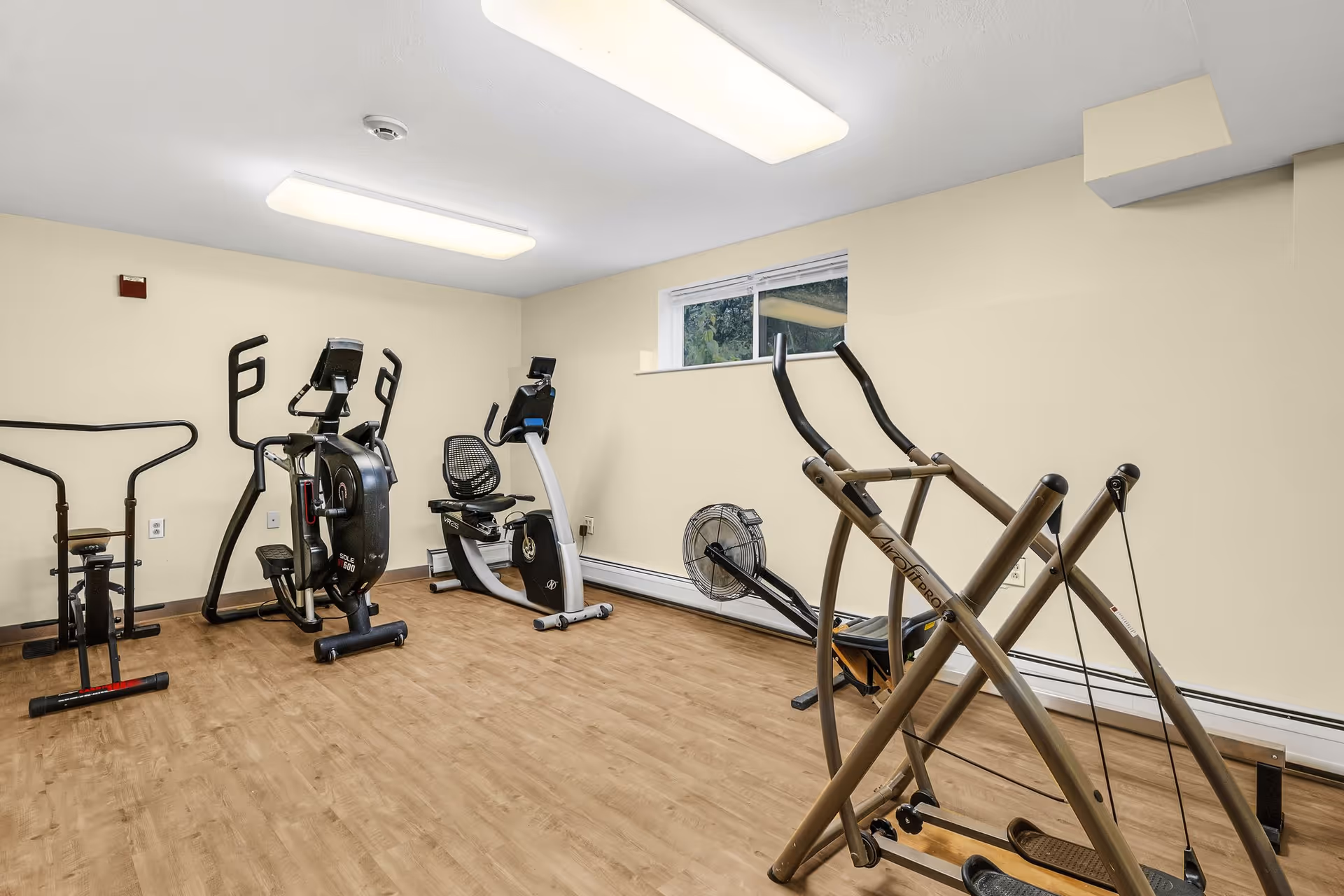 A small fitness room with light beige walls and wood flooring containing various exercise equipment including an elliptical machine, a recumbent bike, a rowing machine, and a stepper. There is a small window near the ceiling letting in natural light and two rectangular ceiling lights providing illumination.
