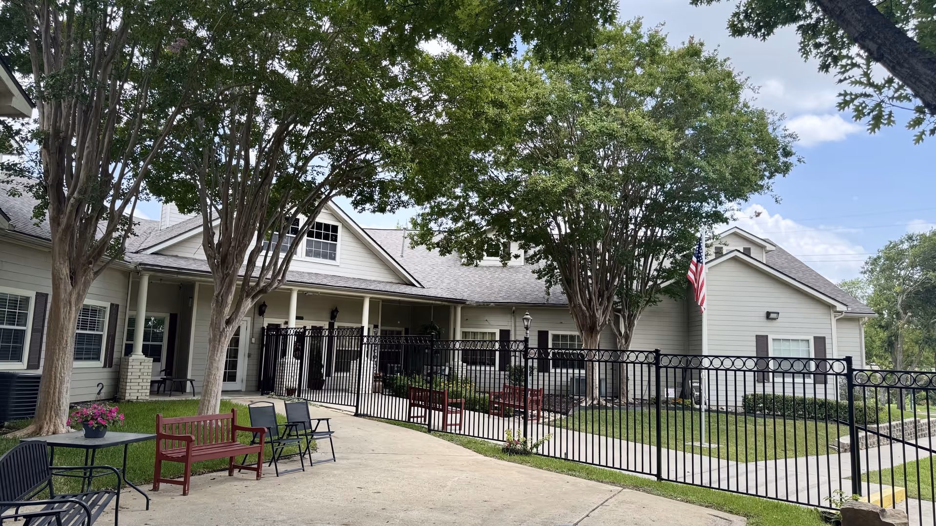 Front exterior of a light-colored senior living building with a fenced courtyard, benches, trees, and an American flag.