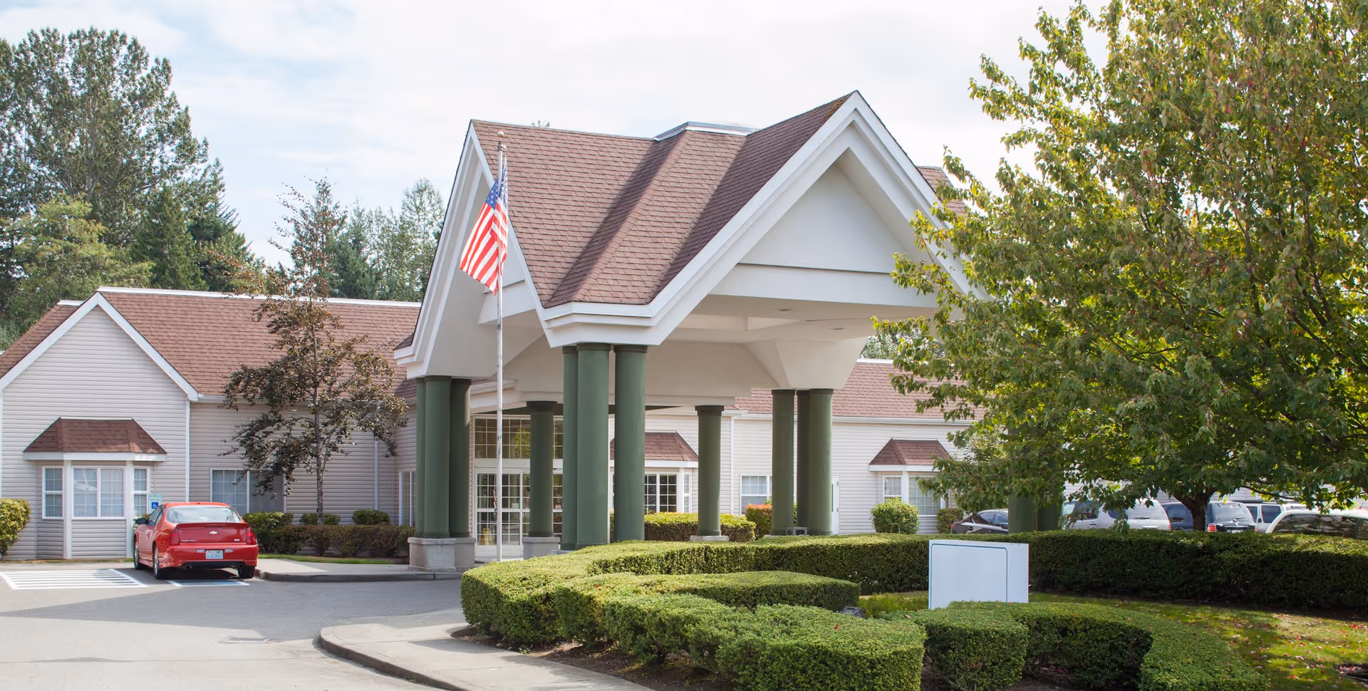 Exterior view of the Everett Center senior living facility showing the entrance with a covered driveway supported by green columns, an American flag, manicured bushes, trees, and a parking area with a red car.