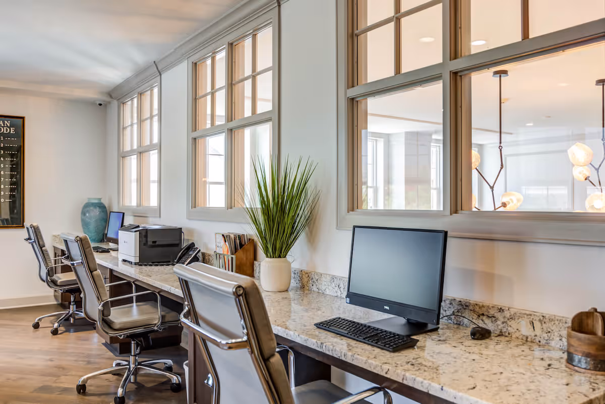 A modern workspace area with a long granite countertop featuring three office chairs, two desktop computers, a printer, a telephone, and decorative items including a tall green plant in a white vase and a large blue vase. The space has large windows with wooden frames allowing natural light to enter.