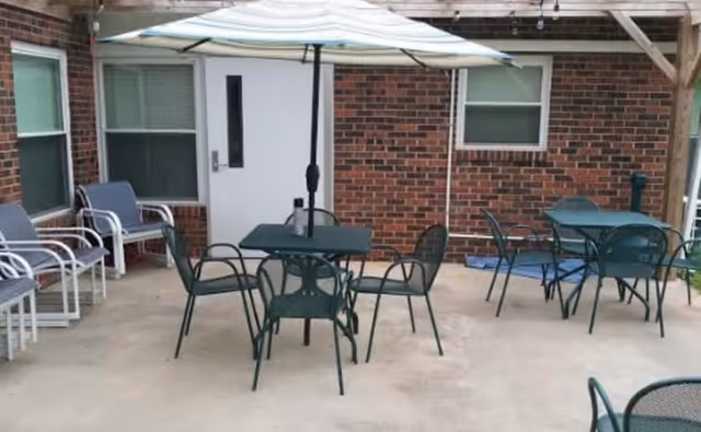 Outdoor patio area with several metal tables and chairs, one table has a large umbrella. The patio is adjacent to a brick building with windows and a white door.