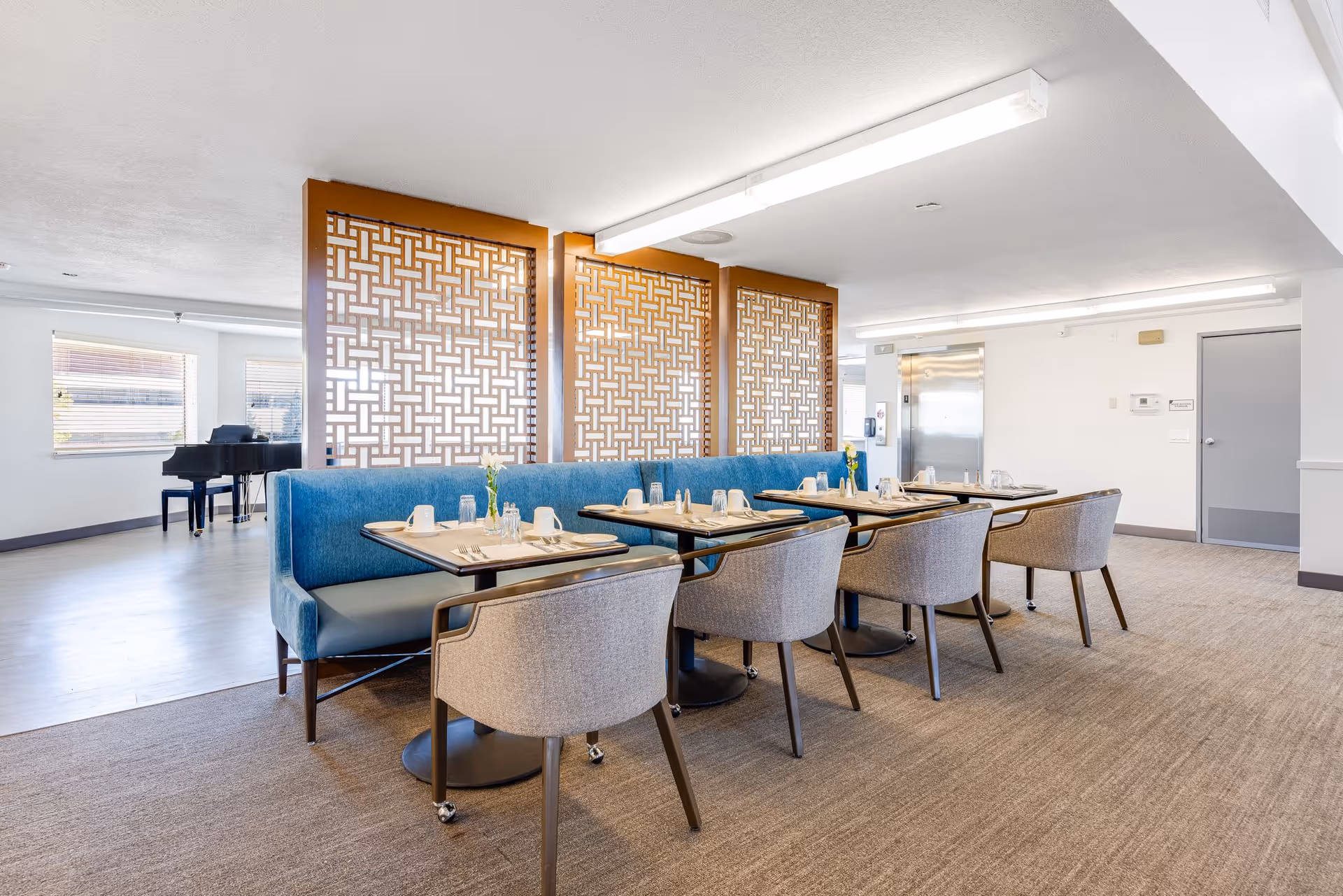 A bright dining area with a row of tables set for meals, featuring blue cushioned bench seating on one side and gray upholstered chairs on the other. Decorative wooden lattice panels separate the dining space from an adjacent area with a black grand piano near windows with blinds. The room has white walls, carpeted flooring in the dining area, and light wood flooring in the adjacent space.