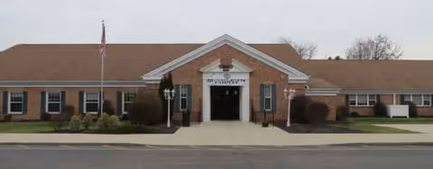 Single-story brick senior living facility with front entrance, flagpole, shrubs, and a driveway.
