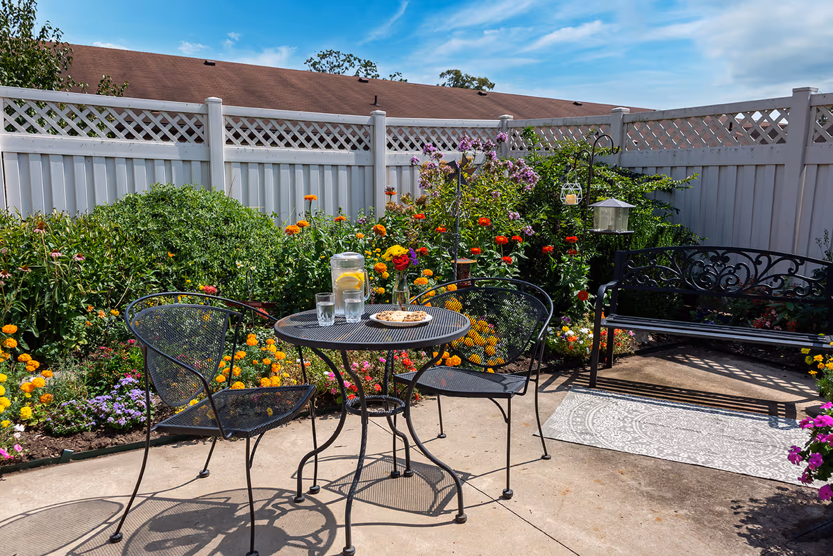 Sunny outdoor patio with a metal table and chairs, a pitcher of lemonade and glasses, a bench, and colorful flower beds against a white fence.
