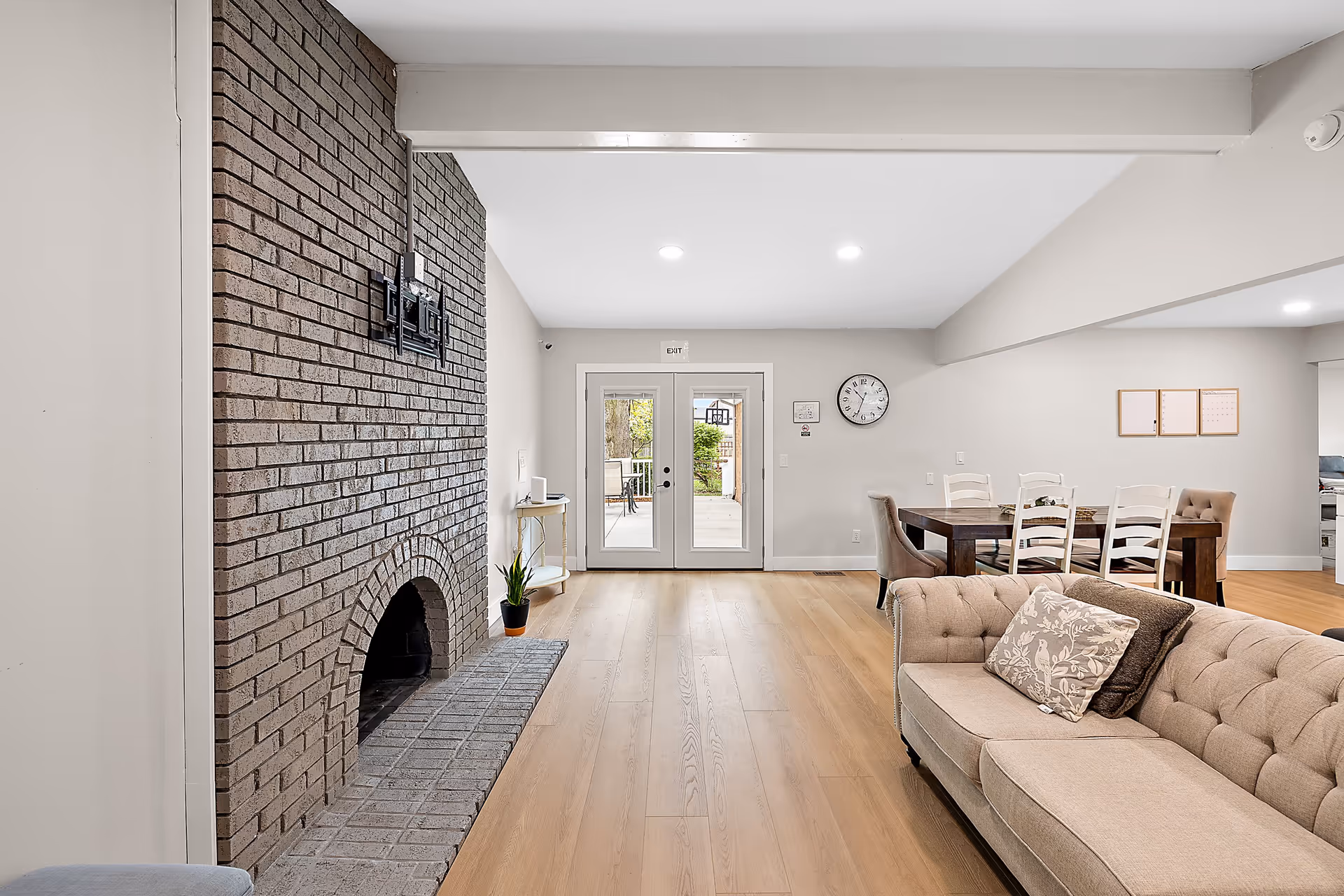 A bright living room with light wood flooring, a beige tufted sofa with decorative pillows, a dark wood dining table with white chairs, and a large brick fireplace on the left wall. Double glass doors lead outside, and a clock and bulletin boards hang on the far wall.
