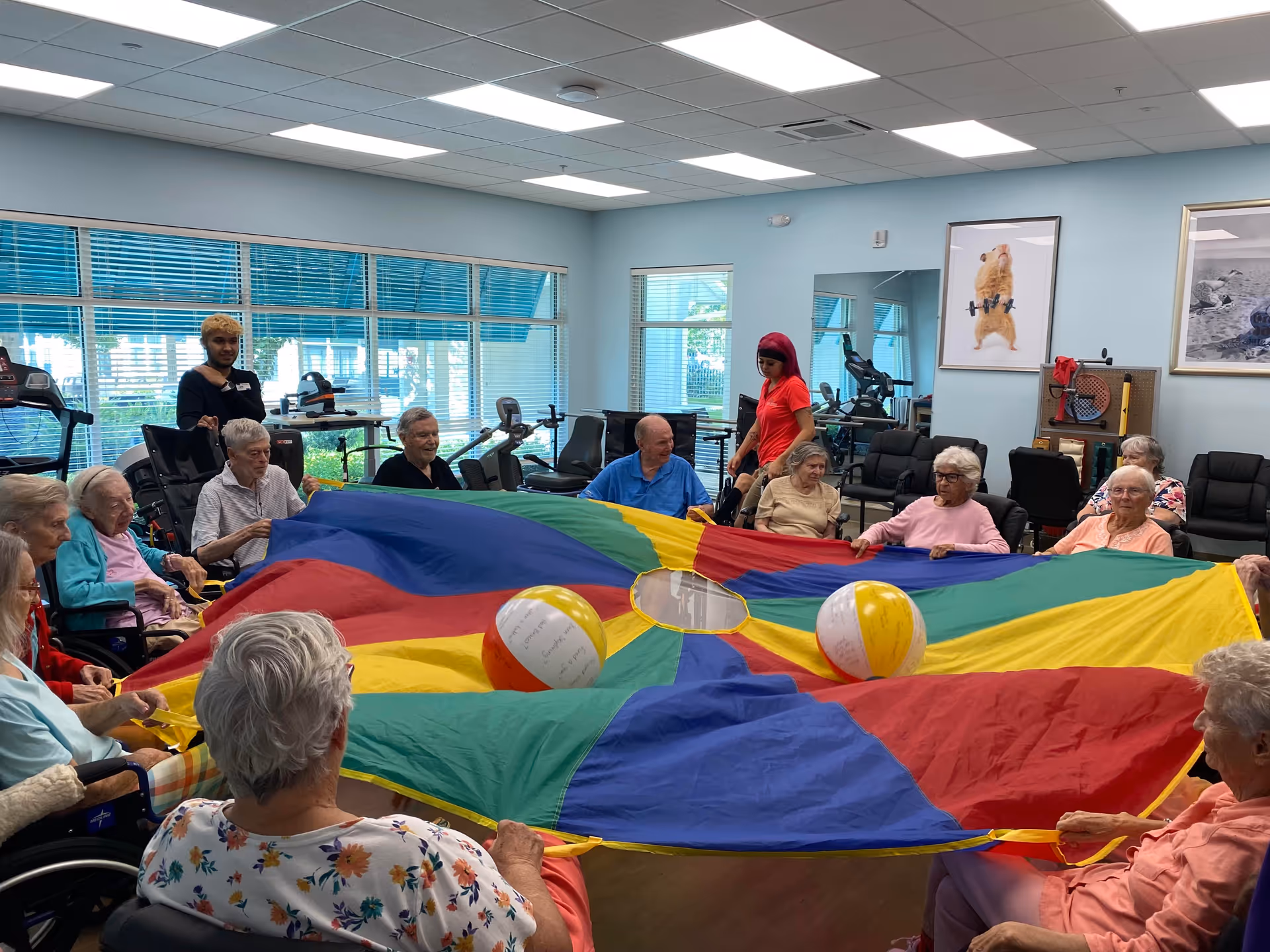 A group of elderly people sitting in a circle in a bright activity room holding the edges of a large colorful parachute with two beach balls on top. Two staff members are standing nearby assisting. The room has large windows, exercise equipment, and framed pictures on the walls.