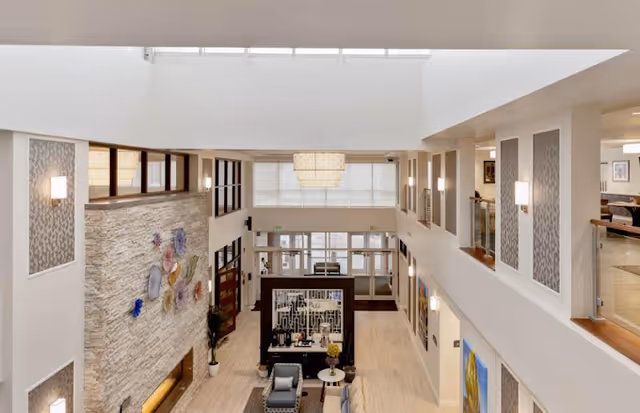 Interior view of a spacious senior living facility lobby with high ceilings and large windows. The area features a stone accent wall with decorative art, comfortable seating arrangements including chairs and a small table, and modern lighting fixtures. There is a mezzanine level with glass railings overlooking the lobby.