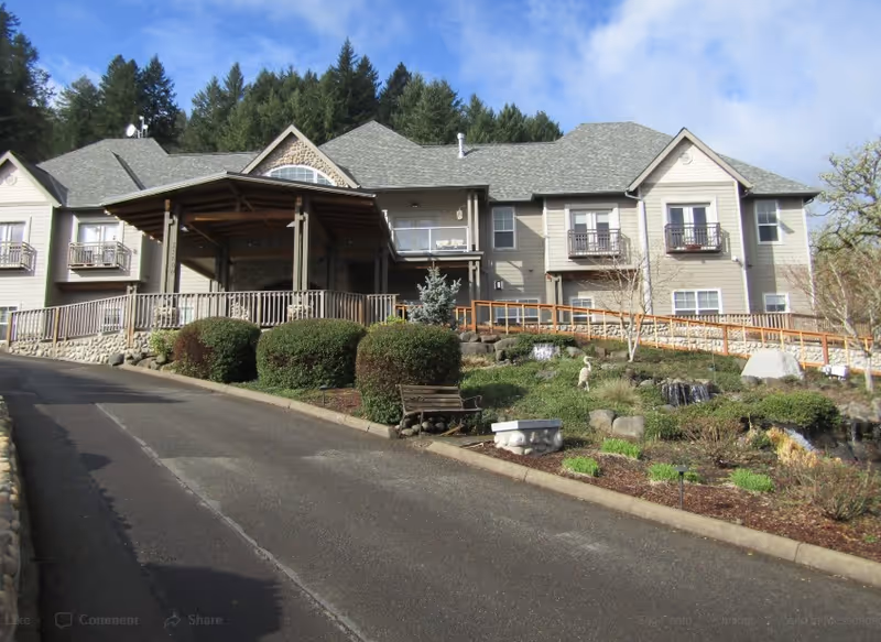 Front exterior of a multi-story retirement community building with a covered entrance, ramp, landscaped grounds and driveway.