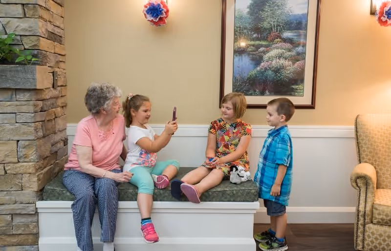 An elderly woman sitting on a cushioned bench next to three children. One girl is holding up a smartphone, showing something to the others. The setting appears to be a cozy indoor area with a stone wall on the left, a framed nature painting on the wall, and a cushioned armchair on the right.