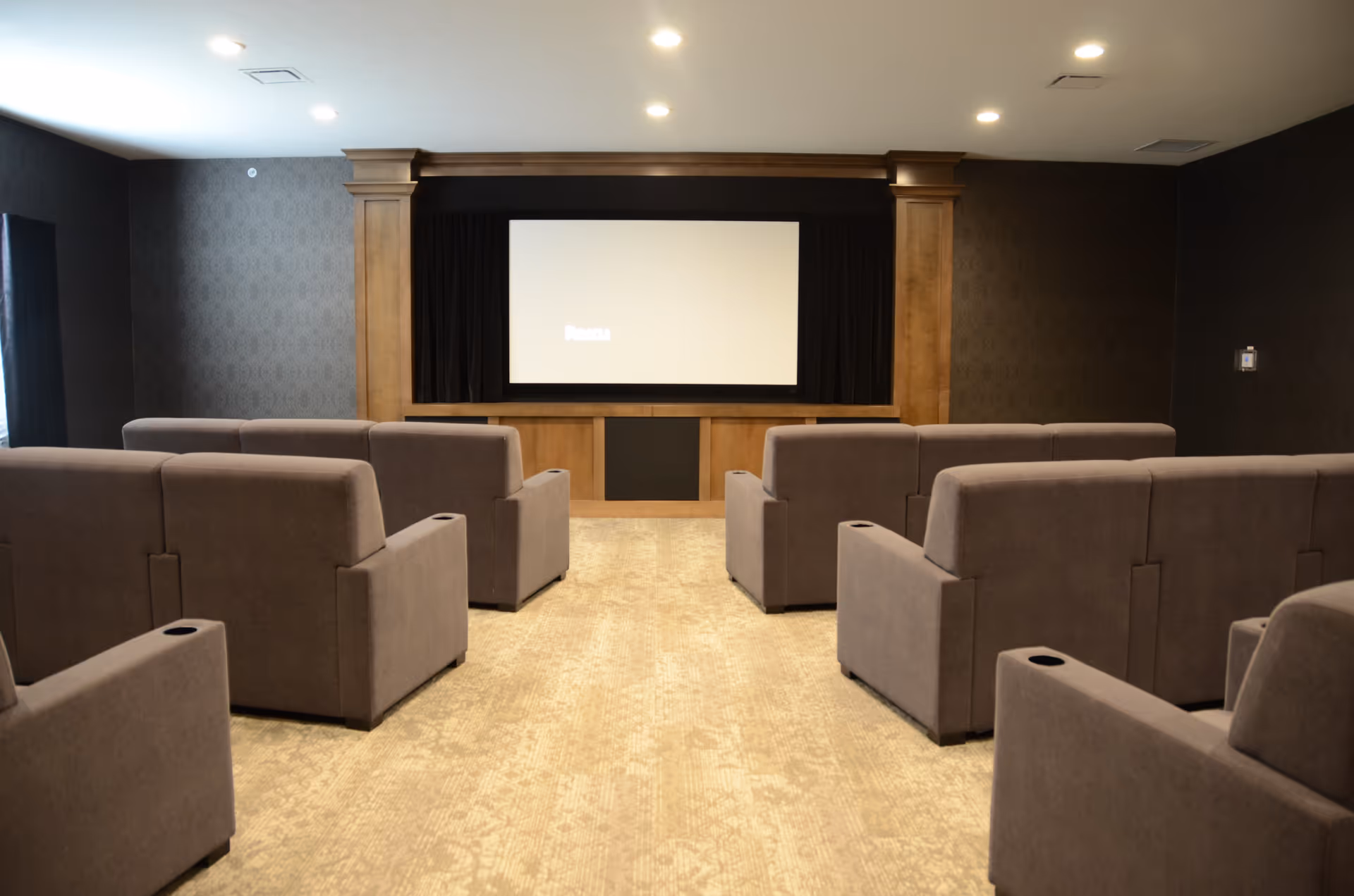 Small screening room with rows of gray reclining chairs facing a projection screen set in a wooden-framed alcove.