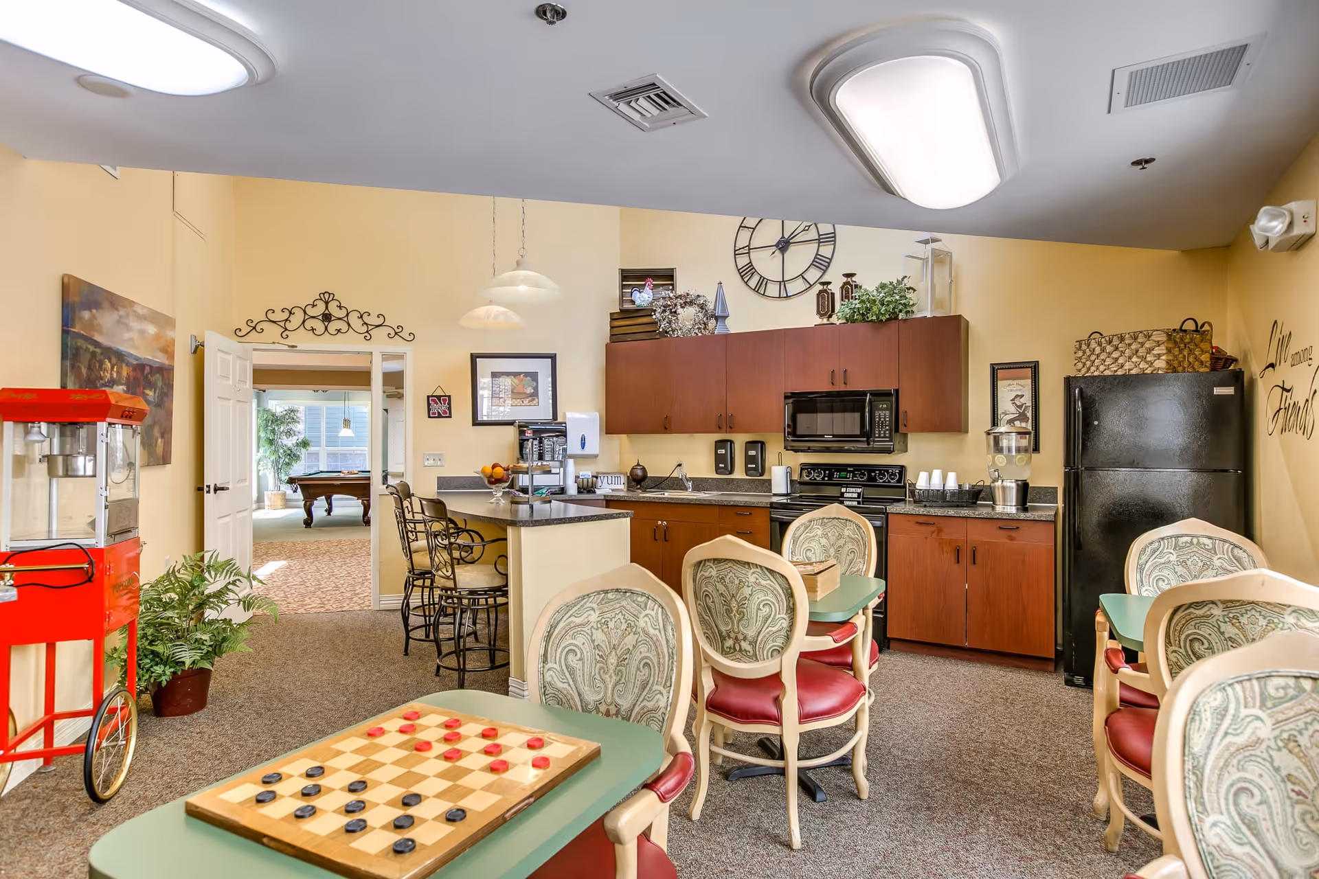 A cozy common area in a retirement community featuring a small kitchen with wooden cabinets, a microwave, stove, and refrigerator. There are several tables with chairs, one table has a checkers board set up. A red popcorn machine is visible on the left side. The walls are painted yellow with decorative artwork and a large clock above the kitchen cabinets. The room is well-lit with ceiling lights and has a carpeted floor. In the background, an open doorway leads to another room with a pool table.