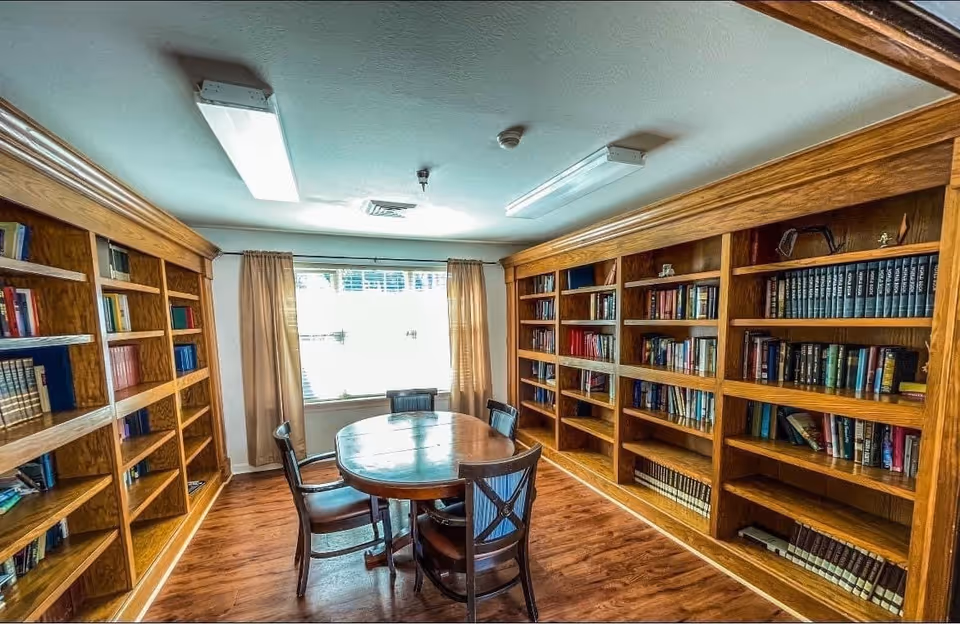 A cozy library room with wooden bookshelves filled with books lining two walls. In the center, there is a round wooden table surrounded by four chairs. A window with beige curtains allows natural light to brighten the room, which has wooden flooring and ceiling lights.