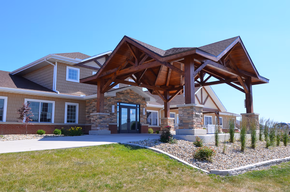 Exterior view of Snyder Village Assisted Living facility showing the main entrance with a large wooden canopy supported by stone pillars, surrounded by landscaped grass and plants under a clear blue sky.