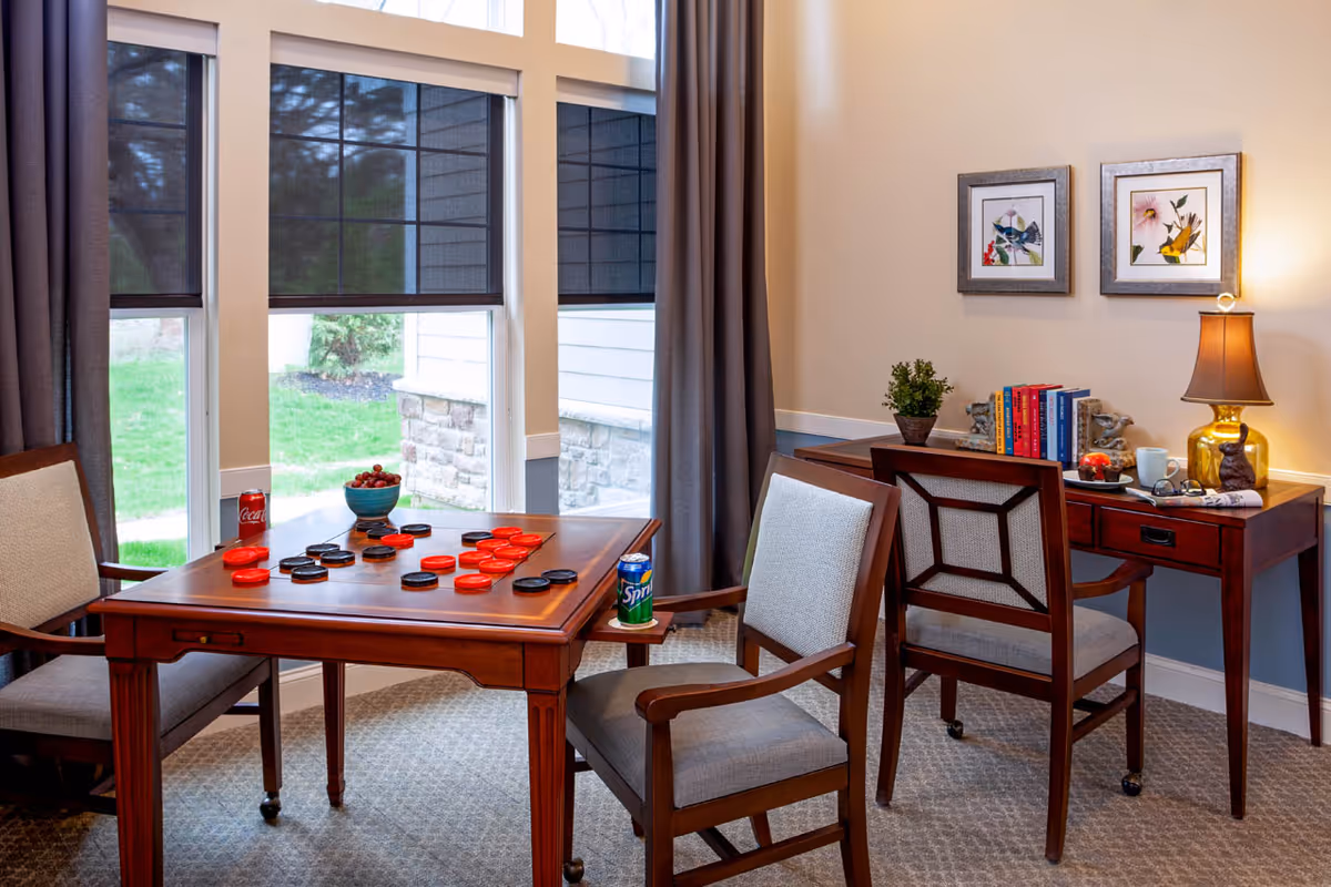 A cozy interior room with a wooden table set for a game of checkers, featuring red and black checkers pieces. Two cushioned wooden chairs are positioned at the table, each with a can of soda (Coca-Cola and Sprite) on the armrest. Behind the table is a wooden desk with a small potted plant, a stack of books, a lamp with a brown shade, and framed bird artwork on the wall. Large windows with dark blinds and curtains allow natural light to enter, showing a green lawn outside.