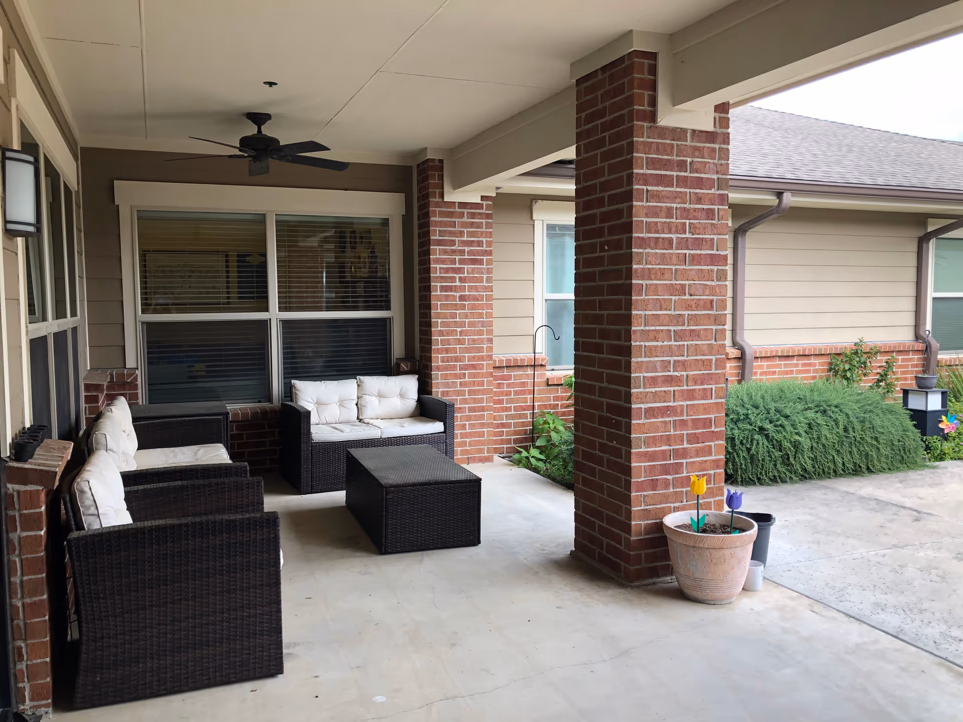 Covered outdoor patio area with dark wicker furniture including two armchairs and a loveseat with white cushions, a matching coffee table, brick pillars, a ceiling fan, and potted plants near the edge of the patio.