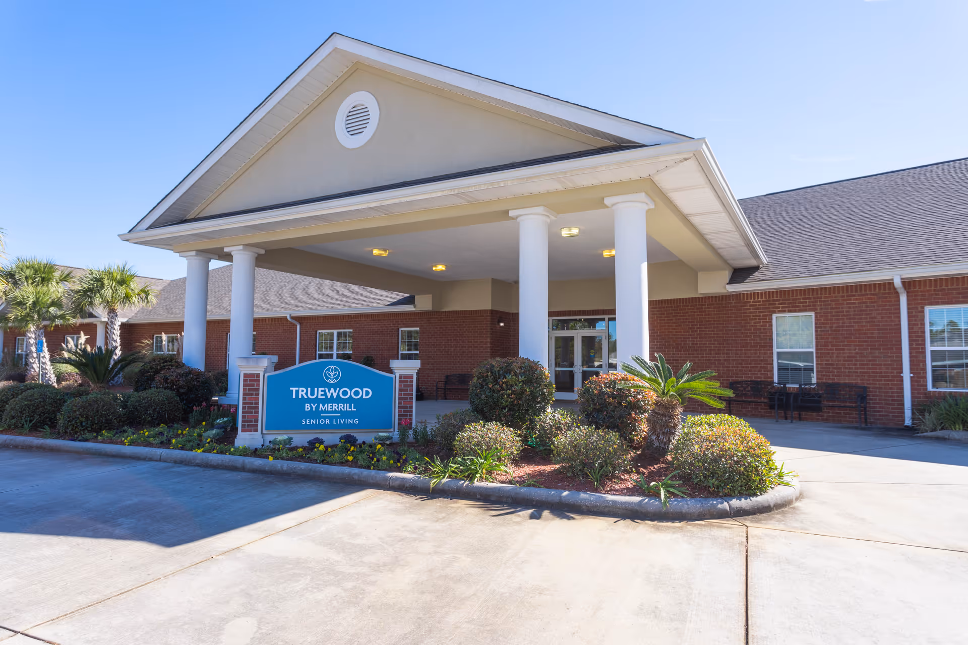 Exterior view of Truewood by Merrill senior living facility in Ocean Springs, showing the entrance with a covered driveway supported by white columns, a blue sign with the facility name, and landscaped bushes and palm trees.