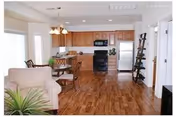 Open-concept living area with a beige armchair and a green plant in the foreground, a wooden dining table with chairs in the middle, and a kitchen with wooden cabinets, black appliances, and a stainless steel refrigerator in the background. The floor is hardwood and the walls are white.