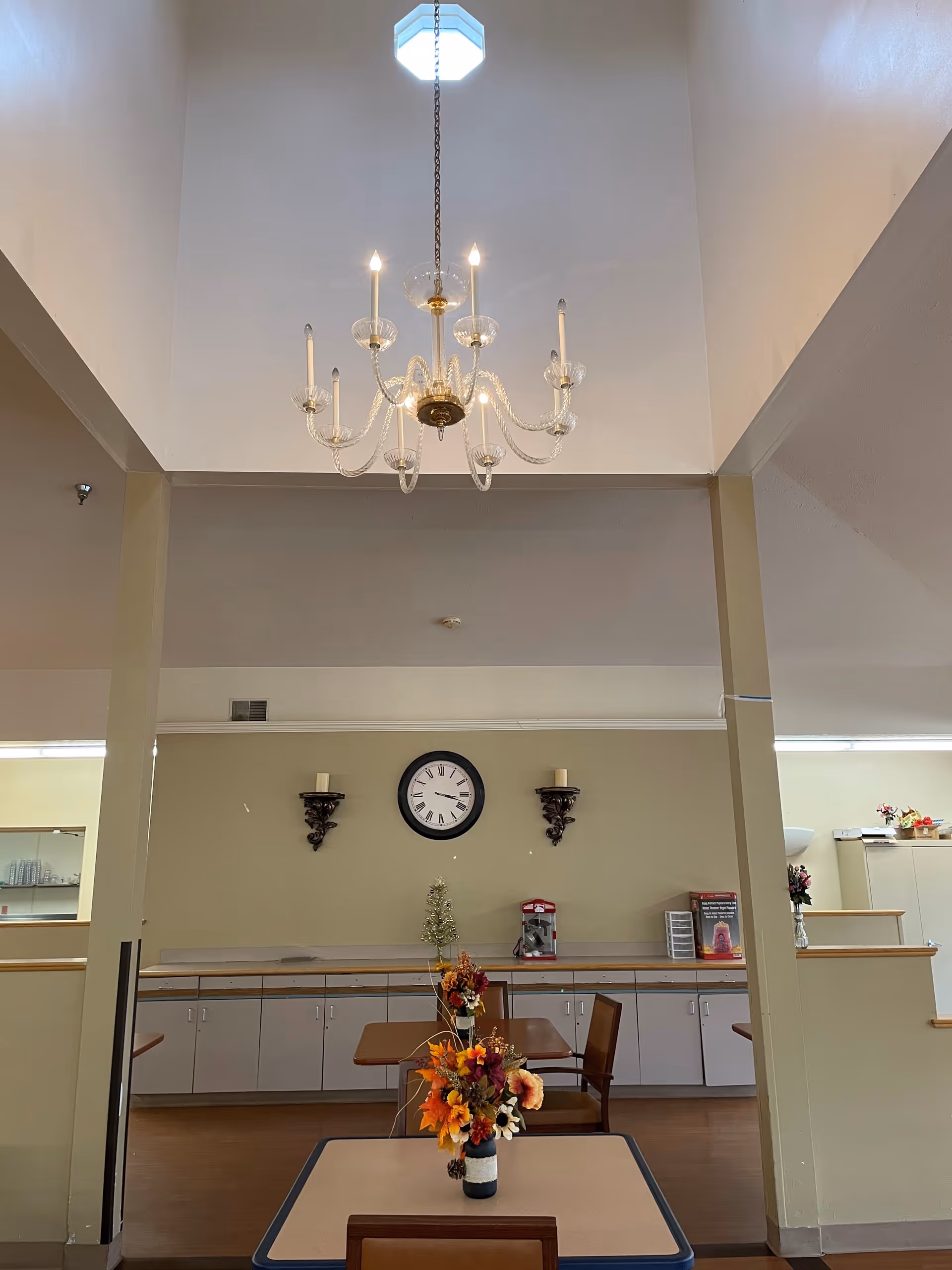Interior view of a common area with a high ceiling and a chandelier hanging from it. Below, there are tables with flower arrangements, a wall clock, and decorative candle holders mounted on the wall. Cabinets and a counter line the back wall, with some items placed on the counter.